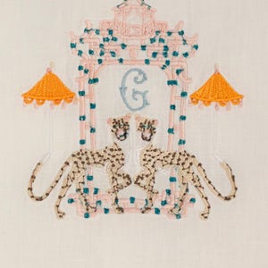 Op de afbeelding: Geborduurde witte linnen theedoek met een roze en turquoise pagode-ontwerp, twee cheetah-figuren en een lichtblauwe letter G. De pagode heeft twee oranje parasols aan weerszijden.