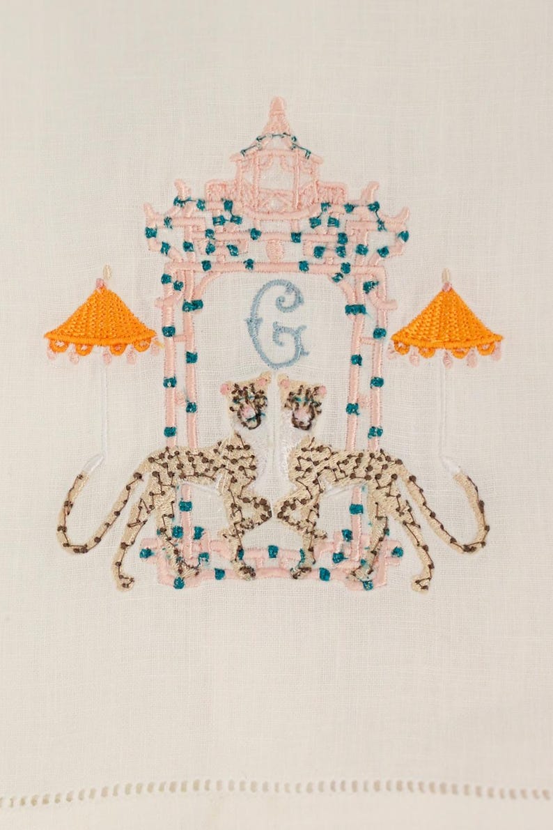 Op de afbeelding: Geborduurde witte linnen theedoek met een roze en turquoise pagode-ontwerp, twee cheetah-figuren en een lichtblauwe letter G. De pagode heeft twee oranje parasols aan weerszijden.