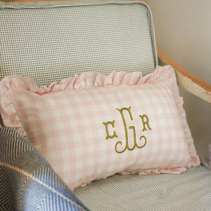 May include: A pink and white gingham cushion with a frilled edge and gold embroidered initials sits on a chair with a chequered pattern. A blue and white patterned blanket is partially visible.