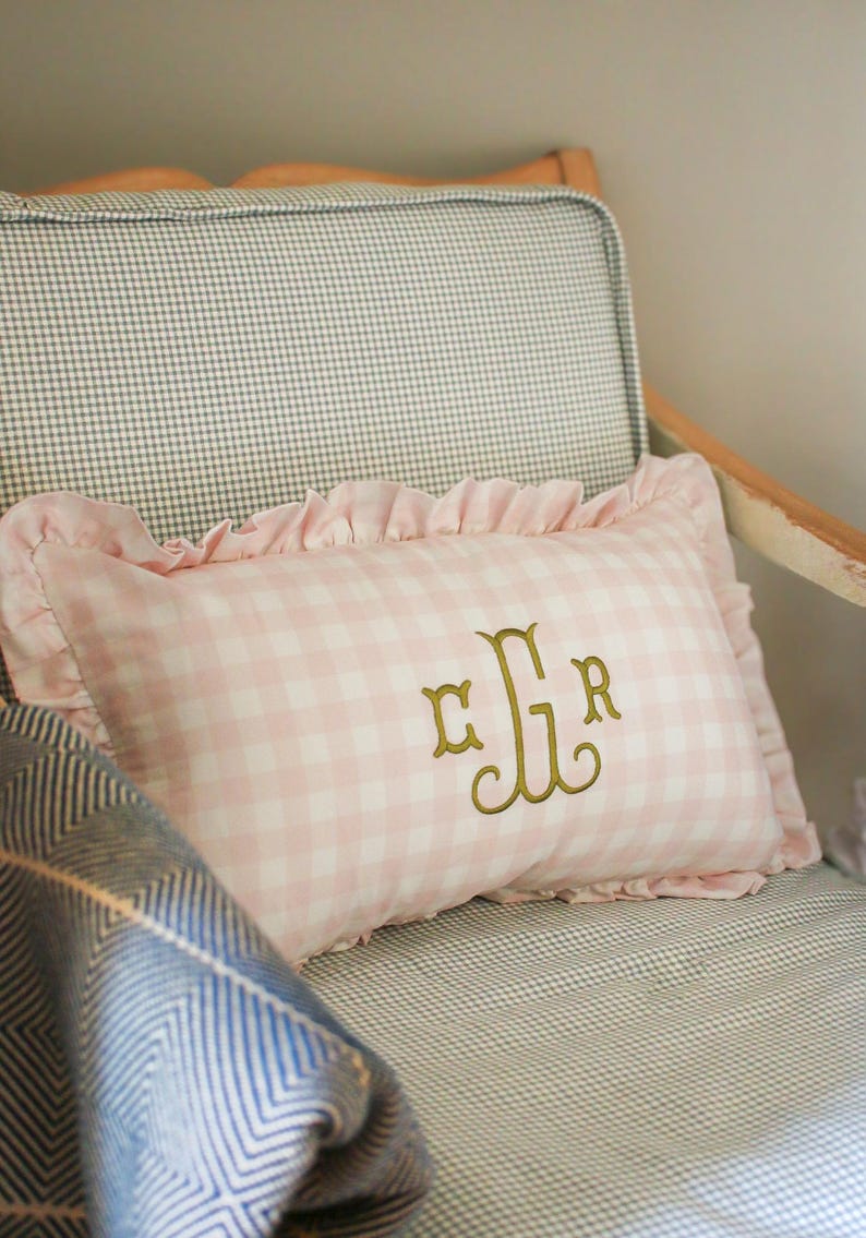May include: A pink and white gingham cushion with a frilled edge and gold embroidered initials sits on a chair with a chequered pattern. A blue and white patterned blanket is partially visible.