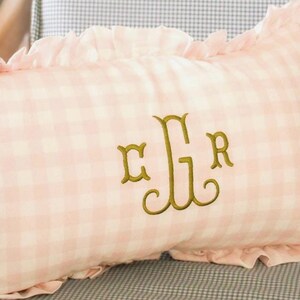 May include: A rectangular pink gingham cushion with a frilled edge. The cushion features an embroidered monogram in gold thread. The cushion has a light pink and white checkered pattern.