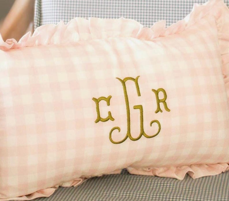 May include: A rectangular pink gingham cushion with a frilled edge. The cushion features an embroidered monogram in gold thread. The cushion has a light pink and white checkered pattern.