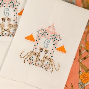 Op de afbeelding: Witte handdoek met geborduurde roze en blauwe pagode met een 'G'-monogram en twee luipaarden. De handdoek ligt op een roze en groene bloemenstof.