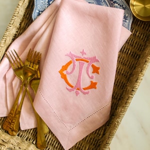 Könnte beinhalten: Ein rosa Leinen-Servietten mit einem gestickten Monogramm in Orange und Rosa, präsentiert in einem Weidenkorb mit goldenen Gabeln. Die Serviette hat einen genähten Rand. Eine kleine goldene Schüssel und ein blau-weiß gemustertes Geschirr sind ebenfalls im Korb.