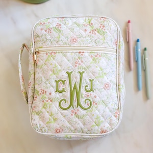 May include: Quilted floral print bag with a gold zip and handle. The bag features a green embroidered monogram and is accompanied by three pens in pink, blue, and green. The bag is approximately 20 cm wide and 15 cm tall.