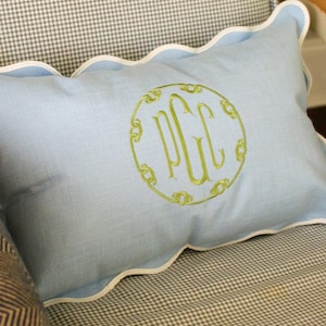 May include: A pale blue rectangular cushion with a scalloped edge and white trim. It has a green monogram in a circular design. The cushion is on a chair with a grey and white check pattern.