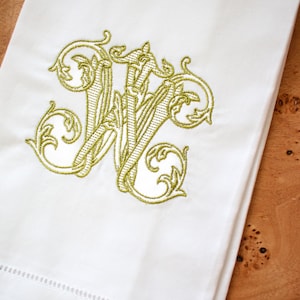 K&ouml;nnte beinhalten: Wei&szlig;es Leinen-Handtuch mit einem goldenen gestickten Monogramm des Buchstabens "W" in einer dekorativen Schriftart.