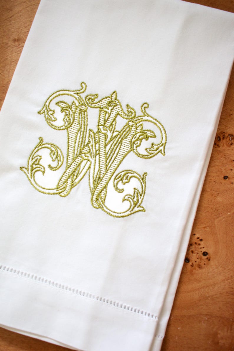 K&ouml;nnte beinhalten: Wei&szlig;es Leinen-Handtuch mit einem goldenen gestickten Monogramm des Buchstabens "W" in einer dekorativen Schriftart.