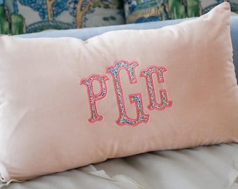 May include: A rectangular blush pink cushion embroidered with the initials PGC in a floral design. The letters are outlined in pink thread. The cushion is on a bed with other cushions in the background.