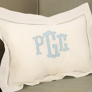 May include: A rectangular off-white pillowcase featuring a wide flange border with delicate hemstitch detailing. The centre of the pillowcase is embroidered with a large, light blue monogram featuring the letters P, G, and C in a decorative font.