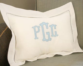 May include: A rectangular off-white pillowcase featuring a wide flange border with delicate hemstitch detailing. The centre of the pillowcase is embroidered with a large, light blue monogram featuring the letters P, G, and C in a decorative font.