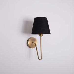 May include: A wall-mounted lamp with a black lampshade and a brass-colored metal frame. The lamp has a curved arm and a circular base. The lampshade is cone-shaped and the lamp is against a white wall.