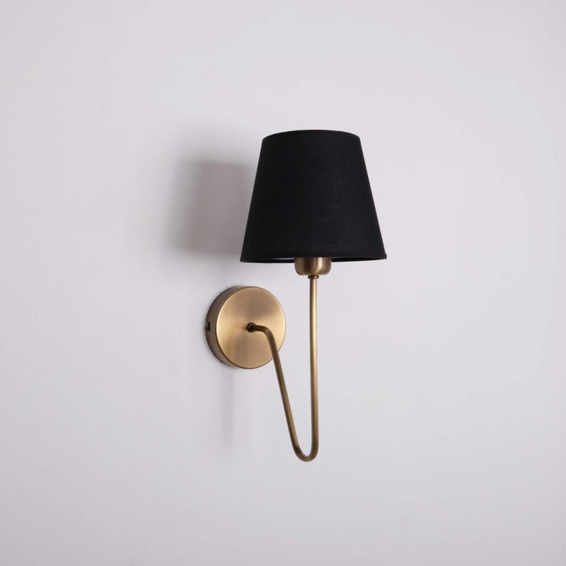 May include: A wall-mounted lamp with a black lampshade and a brass-colored metal frame. The lamp has a curved arm and a circular base. The lampshade is cone-shaped and the lamp is against a white wall.
