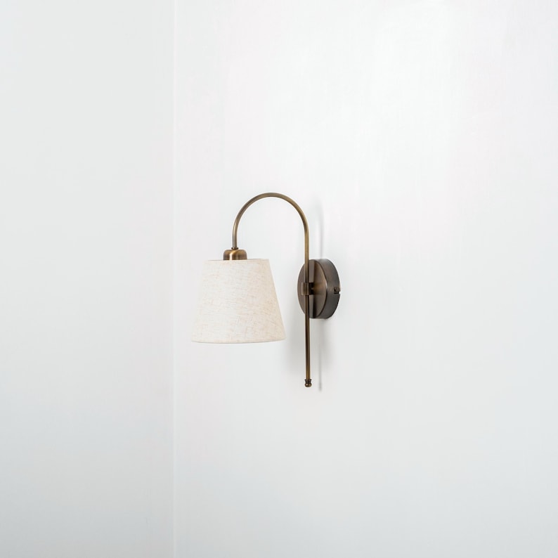Puede incluir: Una l&aacute;mpara de pared de lat&oacute;n con un brazo curvo y una pantalla de tela blanca. La l&aacute;mpara tiene una base redonda con un acabado negro.