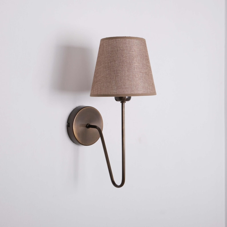 May include: A wall-mounted sconce with a brown lampshade and a bronze-colored metal frame. The lampshade is a tapered cone shape. The sconce is designed to be mounted on a wall and provides ambient lighting.