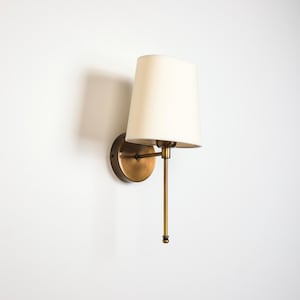 May include: A wall-mounted light fitting featuring a circular brass base, a slender vertical brass arm, and a tapered off-white fabric lampshade. The design includes a horizontal arm connecting the base to the vertical stem and shade.