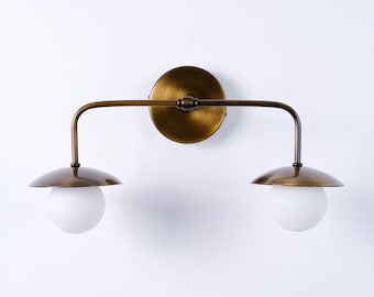 May include: A wall-mounted light fitting featuring a circular brass backplate and a horizontal arm extending to two downward-facing lights. Each light consists of a polished brass dome shade paired with a spherical, frosted white glass globe.