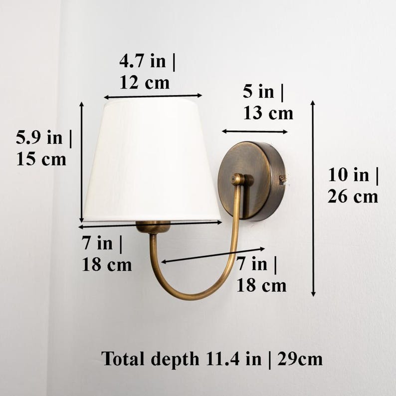 May include: A wall-mounted sconce with a white lampshade and a brass-colored metal frame. The sconce is 10 inches (26 cm) tall, 7 inches (18 cm) wide, and has a total depth of 11.4 inches (29 cm). The lampshade is 4.7 inches (12 cm) wide.