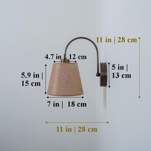 May include: A wall-mounted light fixture with a curved arm and a beige fabric lampshade. The fixture measures 11 inches in height and 5 inches in width. The lampshade is 7 inches in diameter and 4.7 inches in height.