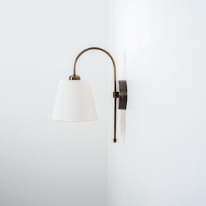 Puede incluir: Una l&aacute;mpara de pared de lat&oacute;n con una pantalla de tela blanca. La l&aacute;mpara tiene un brazo curvo y una base redonda.