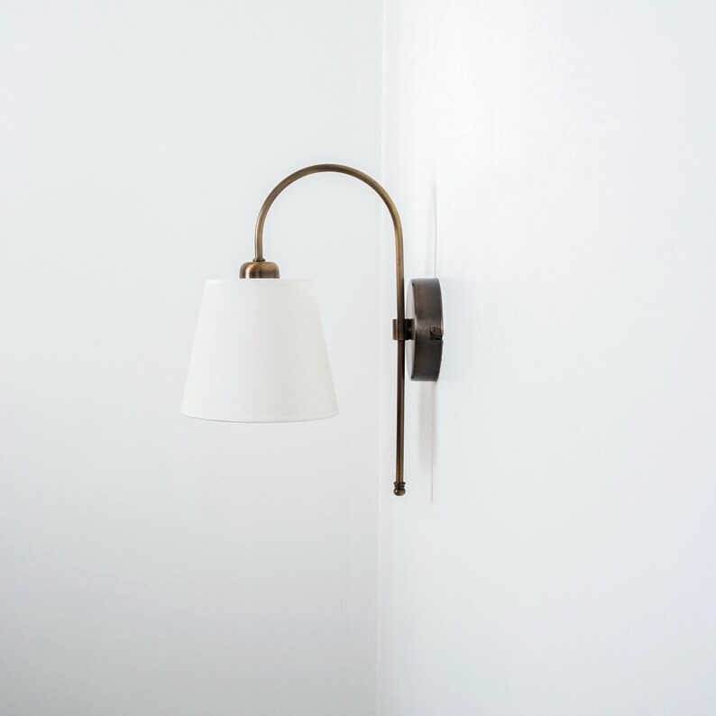 Puede incluir: Una l&aacute;mpara de pared de lat&oacute;n con una pantalla de tela blanca. La l&aacute;mpara tiene un brazo curvo y una base redonda.