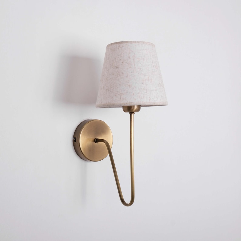 May include: A wall-mounted lamp with a beige fabric shade and a brass-colored metal frame. The lamp has a circular base and a curved arm supporting the shade. The shade is cone-shaped.