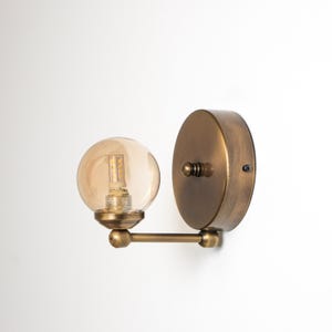 Op de afbeelding: Een wandlamp met een ronde voet van antiek messing en een horizontale arm die uitloopt in een bolvormige, amberkleurige glazen kap. De kap bevat een zichtbare ledlamp en de metalen onderdelen hebben een geborstelde afwerking.
