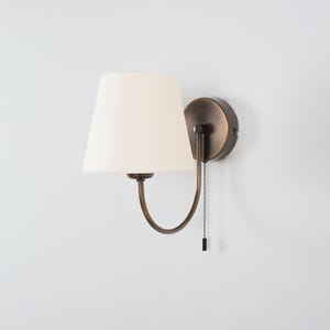 May include: A wall-mounted sconce with a cream-colored lampshade and a bronze-toned metal frame. The sconce has a pull chain for easy operation. The design is classic and suitable for various interior styles.