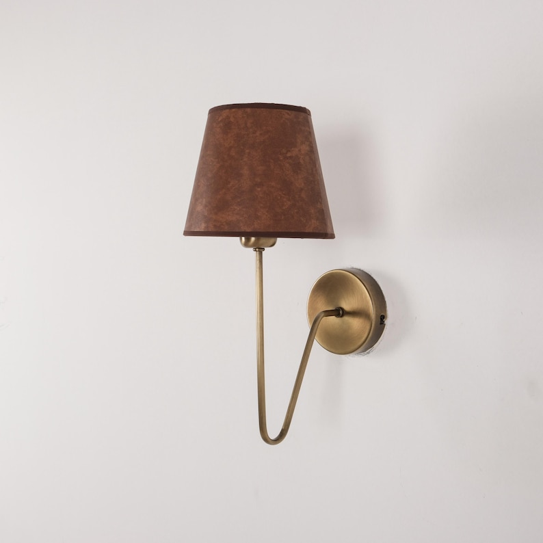 May include: A wall-mounted lamp with a brown lampshade and a brass-colored metal frame. The lampshade is a tapered cone shape. The lamp is mounted on a white wall.