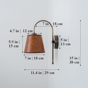 Puede incluir: Una l&aacute;mpara de pared de lat&oacute;n con una pantalla de cuero marr&oacute;n. La l&aacute;mpara mide 38 cm de alto y tiene un brazo de 18 cm. La pantalla tiene un di&aacute;metro de 15 cm y una altura de 12 cm.