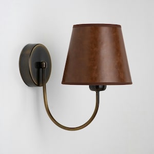 Può includere: Una lampada da parete con paralume marrone e braccio curvo color ottone. La lampada ha una base color bronzo scuro e un paralume marrone con finiture abbinate. Il design è semplice ed elegante.