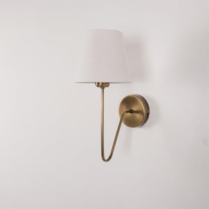 May include: A wall-mounted lamp with a white lampshade and a brass-colored metal frame. The lamp has a curved arm that connects the shade to a circular base. The shade is a light, neutral color, and the lamp is against a white wall.