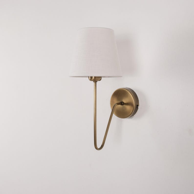 May include: A wall-mounted lamp with a white lampshade and a brass-colored metal frame. The lamp has a curved arm that connects the shade to a circular base. The shade is a light, neutral color, and the lamp is against a white wall.