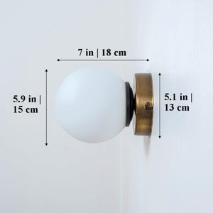 May include: A white frosted glass globe wall sconce with a brass base. The sconce measures 7 inches wide, 5.9 inches tall, and 5.1 inches deep.