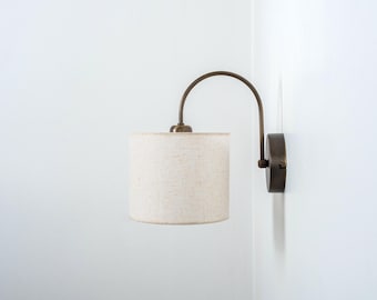 May include: A wall-mounted light fixture with a curved arm and a white fabric shade. The fixture is made of brass and has a simple, modern design.