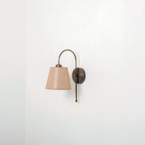 Puede incluir: Una l&aacute;mpara de pared con un brazo curvo y una pantalla de tela beige. La l&aacute;mpara est&aacute; acabada en lat&oacute;n cepillado.