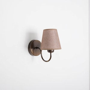 Könnte beinhalten: Eine Wandleuchte aus Bronze mit einem kleinen, konischen Lampenschirm in Beige. Die Leuchte hat einen gebogenen Arm und eine runde Basis. Geeignet für die Innenbeleuchtung.