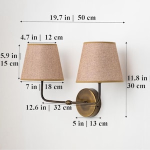 May include: A double-arm wall sconce with two fabric lampshades. The sconce is 50 cm wide, 30 cm tall, and the arms extend 32 cm. The lampshades are 18 cm in diameter and 15 cm tall.