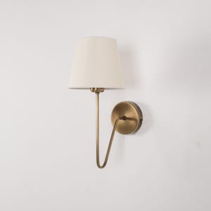 May include: A wall-mounted lamp with a cream-colored lampshade and a brass-toned metal frame. The lamp has a curved arm and a circular base, designed for indoor lighting. The fixture is mounted on a white wall.
