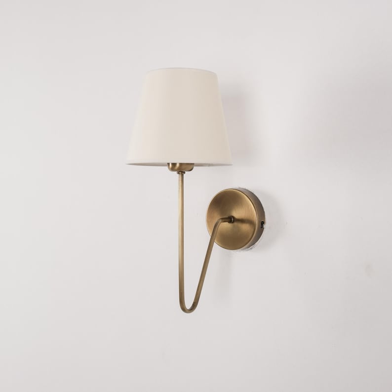 May include: A wall-mounted lamp with a cream-colored lampshade and a brass-toned metal frame. The lamp has a curved arm and a circular base, designed for indoor lighting. The fixture is mounted on a white wall.