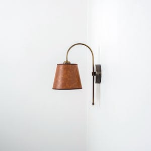 Puede incluir: Una l&aacute;mpara de pared de cuero marr&oacute;n con un brazo curvo y una pantalla de tela marr&oacute;n.