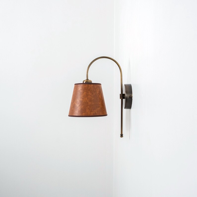 Puede incluir: Una l&aacute;mpara de pared de cuero marr&oacute;n con un brazo curvo y una pantalla de tela marr&oacute;n.