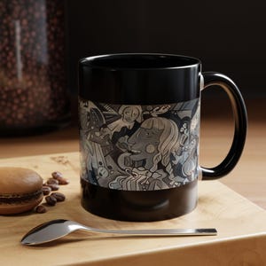 May include: Black ceramic coffee mug with a white and grey abstract design featuring faces and geometric shapes. The mug has a handle on the right side.