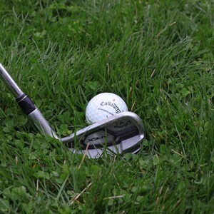 May include: A silver golf club with a black grip resting on green grass next to a white golf ball with the word "Callaway" printed on it.