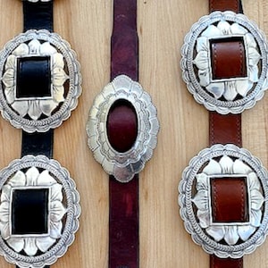 Może przedstawiać: Trzy srebrne klamry do paska z ozdobnymi wzorami kwiatowymi. Klamry są przymocowane do sk&oacute;rzanych pask&oacute;w w kolorze czarnym, brązowym i bordowym.