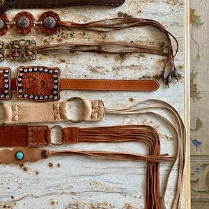 K&ouml;nnte beinhalten: F&uuml;nf Lederg&uuml;rtel mit Fransen. Die G&uuml;rtel sind braun, beige und sandfarben. Einige G&uuml;rtel haben Metallschnallen und dekorative N&auml;hte.