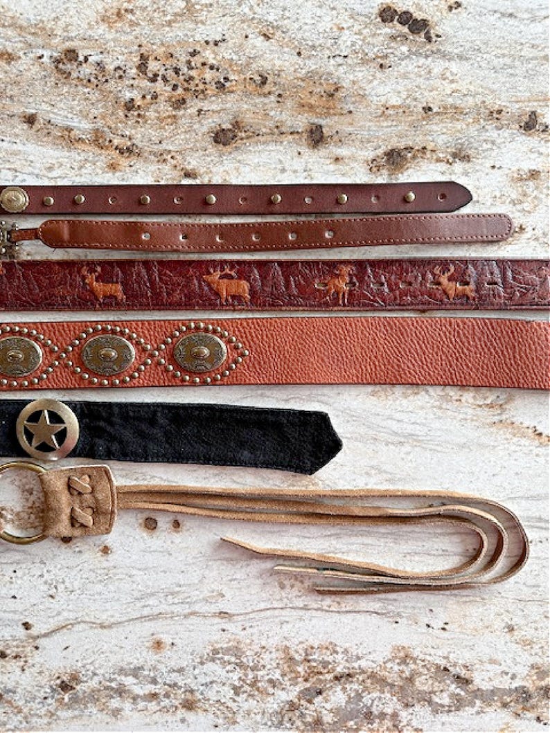 Peut inclure: Cinq ceintures en cuir diff&eacute;rentes avec des motifs et des embellissements vari&eacute;s. Les ceintures sont dispos&eacute;es verticalement sur une surface en bois brun clair. La ceinture du haut est brune avec des clous argent&eacute;s, la deuxi&egrave;me ceinture est brune avec un motif de cerf, la troisi&egrave;me ceinture est orange avec des clous argent&eacute;s et des accents m&eacute;talliques ronds, la quatri&egrave;me ceinture est noire avec un accent m&eacute;tallique en forme d'&eacute;toile, et la cinqui&egrave;me ceinture est brune avec une frange.