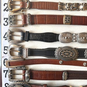 May include: A collection of eight leather belts with various buckle designs. The belts feature different colors, including black and brown, and textures, such as embossed and braided leather. The buckles are made of silver and gold-toned metal.