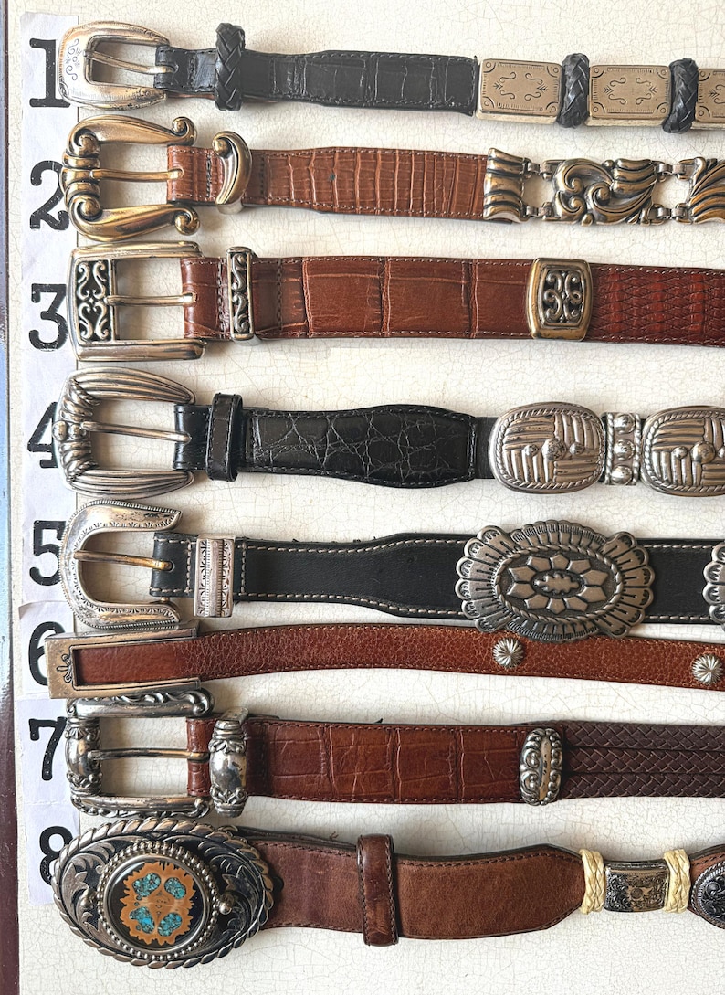 May include: A collection of eight leather belts with various buckle designs. The belts feature different colors, including black and brown, and textures, such as embossed and braided leather. The buckles are made of silver and gold-toned metal.
