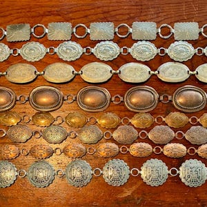 Pode incluir: Uma coleção de cintos de metal vintage com vários designs. Os cintos apresentam elos retangulares, ovais e circulares em tons de prata, ouro e cobre. Os cintos estão dispostos sobre uma superfície de madeira, mostrando seus padrões e texturas intrincados.